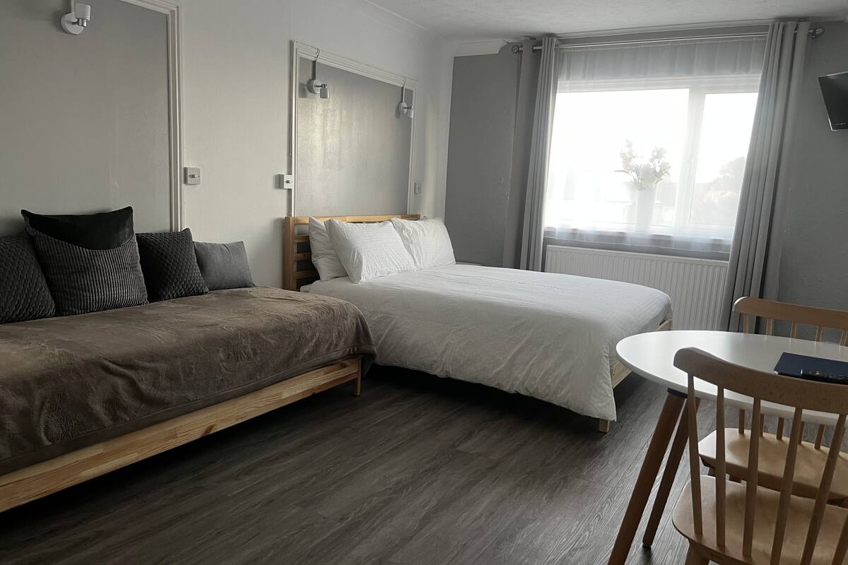 A hotel room with two beds, a wooden floor, a small table with a chair, and a window with curtains.