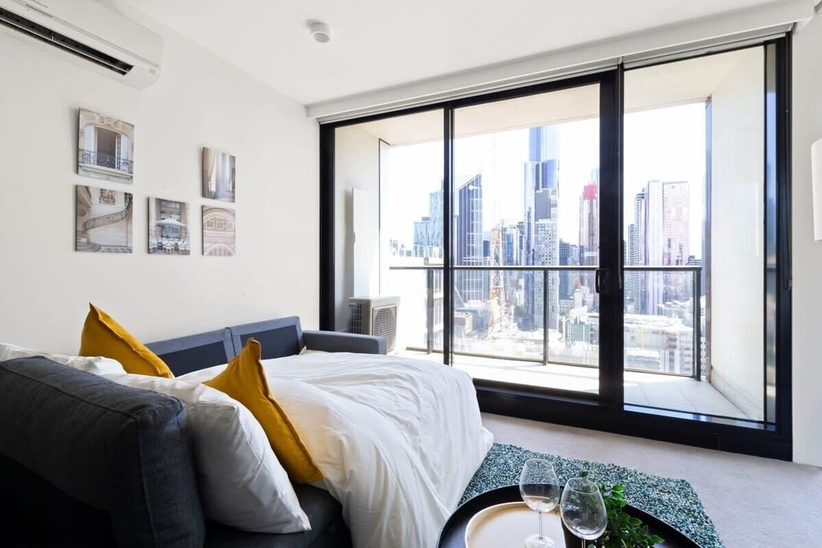 A modern hotel room with a large window offering a city view, a bed with white bedding and yellow pillows, and a small table with glasses.