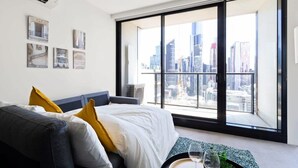 Ein modernes Hotelzimmer mit einem großen Fenster, das einen Blick auf die Stadt bietet, einem Bett mit weißer Bettwäsche und gelben Kissen sowie einem kleinen Tisch mit Gläsern.