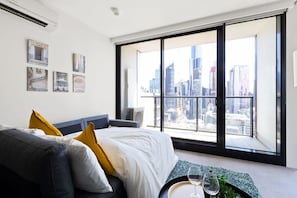 Habitación de hotel moderna con un ventanal grande que ofrece vistas a la ciudad, una cama con ropa blanca y almohadas amarillas, y una mesita con vasos.