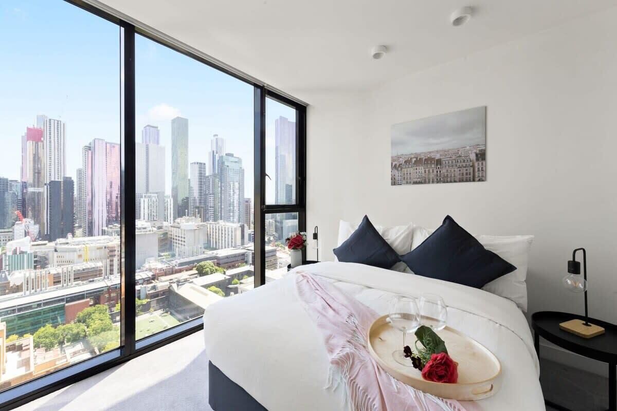 Een moderne hotelkamer met een prachtig stadsgezicht, een keurig opgemaakt bed met een dienblad met wijnglazen en een roos, en een klein nachtkastje met een lamp.