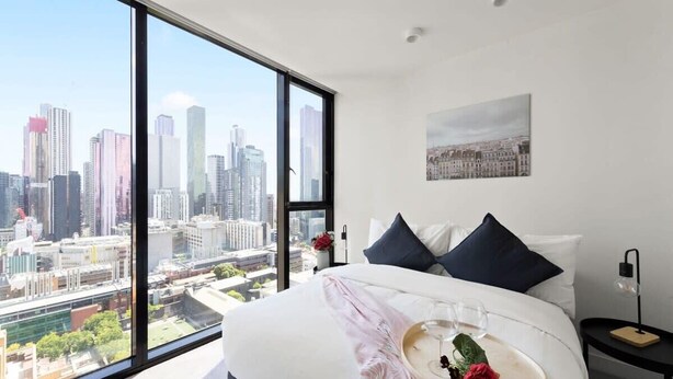 Ein modernes Hotelzimmer mit weitreichendem Stadtblick, einem ordentlich bezogenen Bett, einem Tablett mit Weingläsern und einer Rose sowie ein kleiner Nachttisch mit einer Lampe.