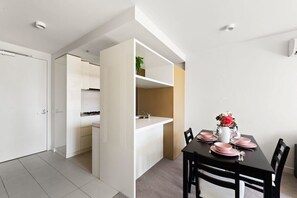 Una cocina moderna con armarios blancos, una zona de comedor con una mesa negra para cuatro personas y una pequeña planta en un estante.