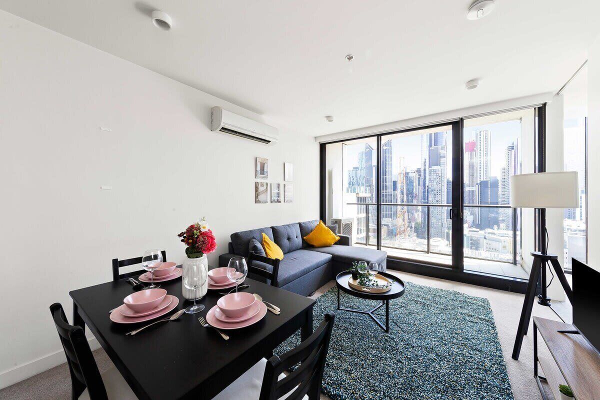 A modern living room with a dining area, large windows with a city view, a gray sofa with yellow cushions, and a black dining table set with pink plates and glasses.