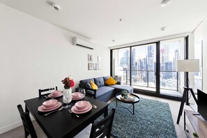 A modern living room with a dining area, large windows with a city view, a gray sofa with yellow cushions, and a black dining table set with pink plates and glasses.