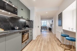 Uma cozinha moderna com forno embutido, micro-ondas e bancada de mármore. É possível ver uma área de jantar com mesa e cadeiras através de uma porta aberta.