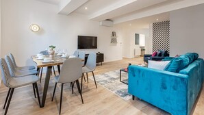 A modern living room with a blue sofa, a dining area with patterned chairs, and a flat-screen TV mounted on the wall.