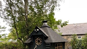 A small, dark wooden structure with a hexagonal window and a chimney, surrounded by a well-maintained lawn and a stone building in the background. - Royal Snug & WEE Snug (Aboyne)