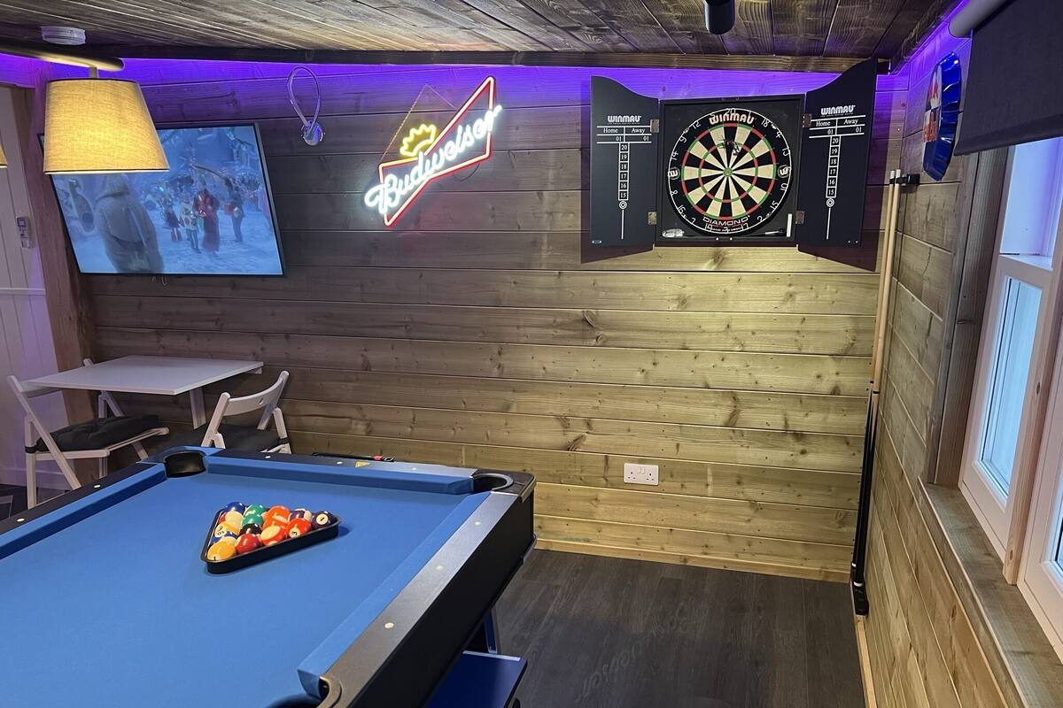 A billiards table with balls, a dartboard, and a neon Budweiser sign in a room with wooden walls and a window.