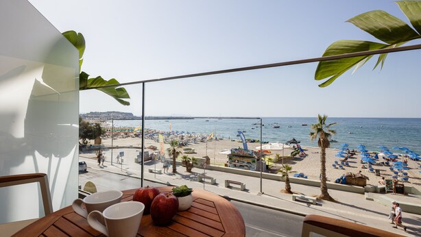 Ein Balkon mit Blick auf einen Strand, ein Holztisch mit Tassen und Äpfeln sowie ein Topfpflanze.