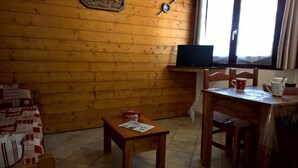 A cozy room with wooden walls, a small table with a newspaper, a TV on a stand, and a window with curtains.