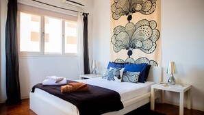 A neatly arranged bedroom with a large bed, a bedside table with a lamp, and a wall with a floral pattern. - Maison Moz - Moldarte (Maputo)