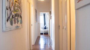 A hallway with wooden flooring and white walls, leading to a room with a bed and a window. - Maison Moz - Moldarte (Maputo)