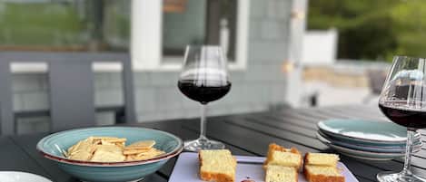 Uma mesa de jantar com diversos alimentos, incluindo pão, queijo e frutas, e dois copos de vinho tinto.