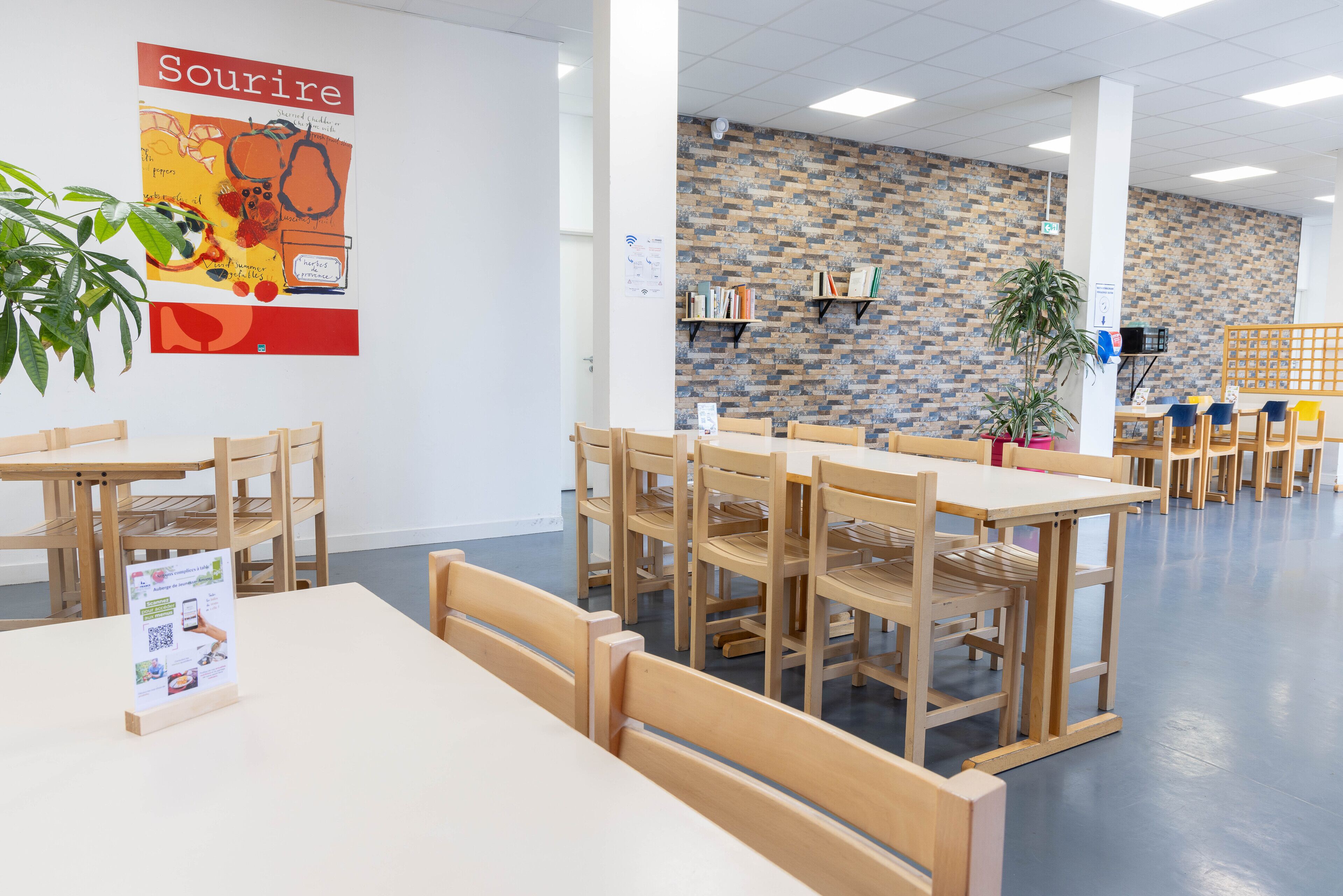 Un espace repas avec des tables et des chaises en bois, un mur décoré d’un motif en briques et une affiche accrochée au mur.