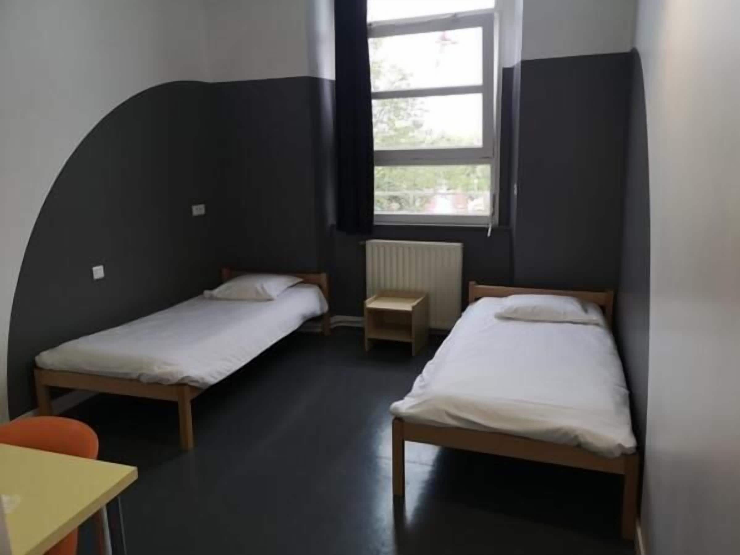 A small, compact room with two single beds, a small table, and a window providing natural light.