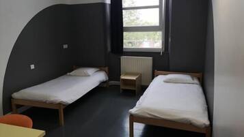 A small, compact room with two single beds, a small table, and a window providing natural light.