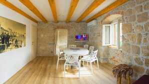Un comedor con una mesa redonda y sillas blancas, paredes de piedra, vigas de madera y un televisor en la pared.