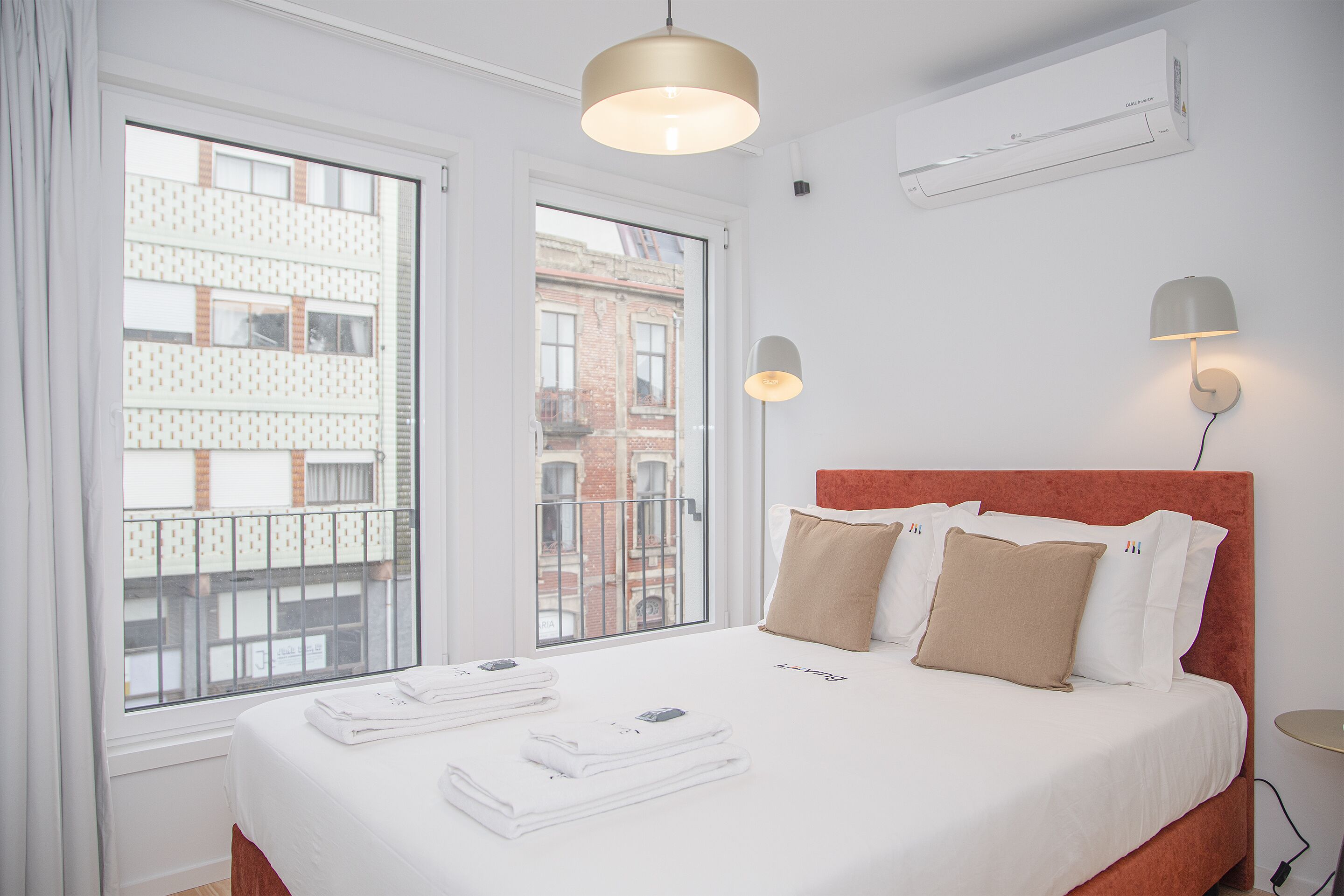 A hotel room with a large bed, two bedside lamps, a window with a view of buildings, and an air conditioning unit on the wall.