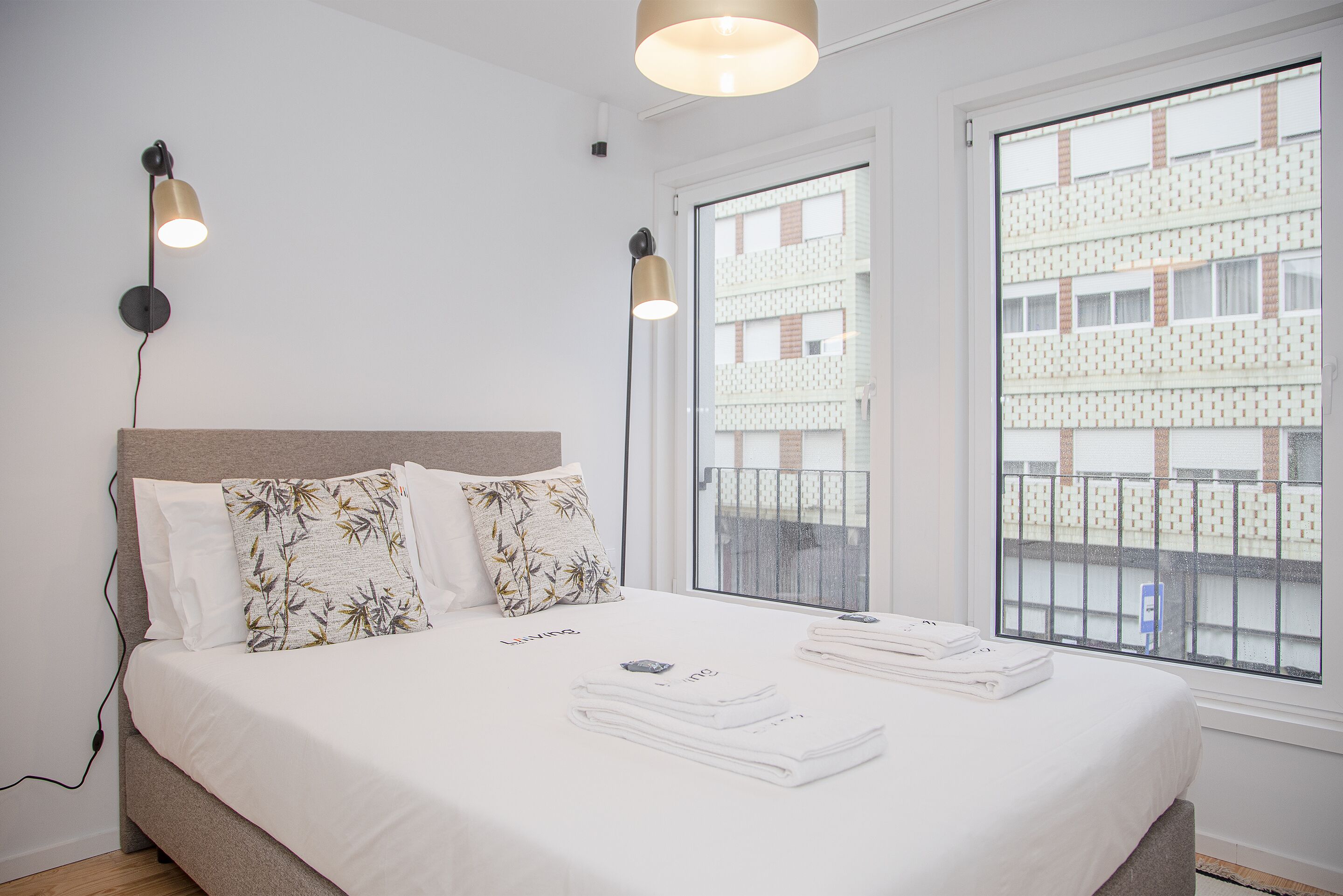 A neatly made bed with white linens and a floral pillow, two bedside lamps, and a view of a building through a window.