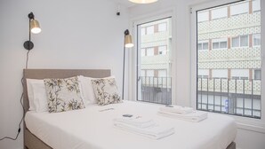 A neatly made bed with white linens and a floral pillow, two bedside lamps, and a view of a building through a window. - Liiiving - Modern & Glam Apartment M (Porto)