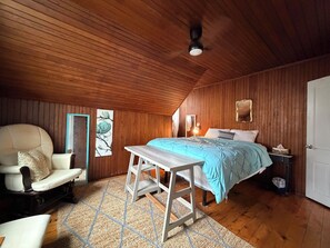Une chambre avec un plafond en bois, un lit recouvert d’une courtepointe bleue, une chaise blanche et un plancher en bois.