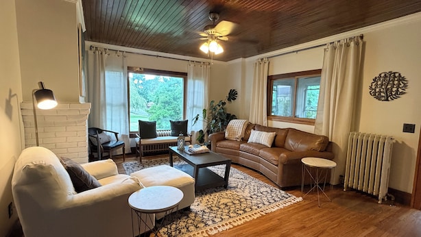 A living room with a sofa, armchair, coffee table, and a fireplace. There are two windows with curtains, a ceiling fan, and a radiator. - Beautiful Lake home-5 minute walk to the Public Beach Area, (Rochester)