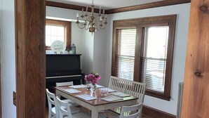 A dining area with a wooden table, white chairs, a chandelier, and a piano in the background. - Beautiful Lake home-5 minute walk to the Public Beach Area, (Rochester)