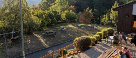 An outdoor dining area with tables and chairs, surrounded by greenery and mountains in the background.