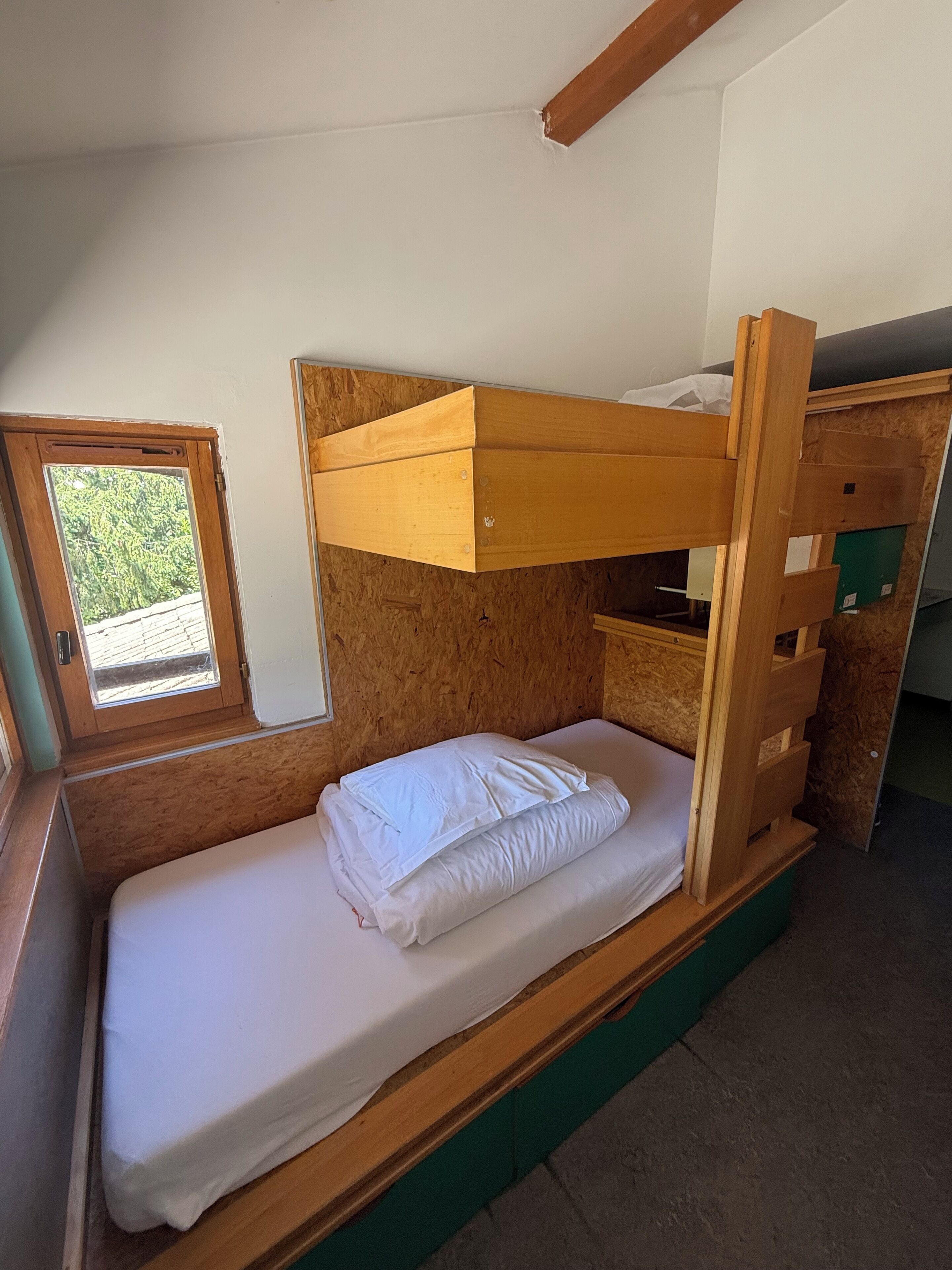 Una cama litera con estructura de madera y ropa de cama blanca, una ventana pequeña con vista a follaje, y una pared verde.