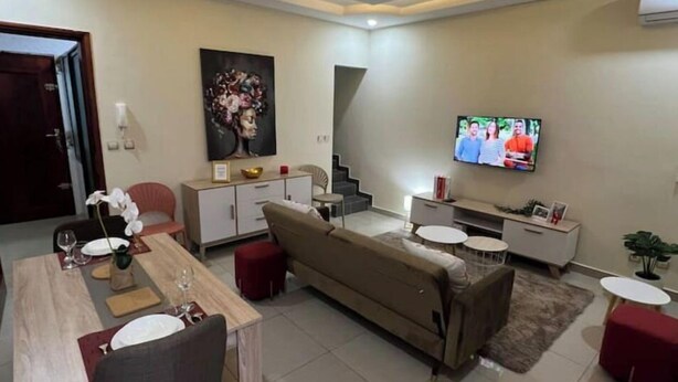 A modern living room with a dining area, a sofa, a TV, and a staircase. - Appart Cozy Riviera 4 Verdant (Abidjan)