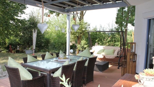 Een terras buiten met rieten meubilair, een tafel met glazen blad en een schommelstoel.