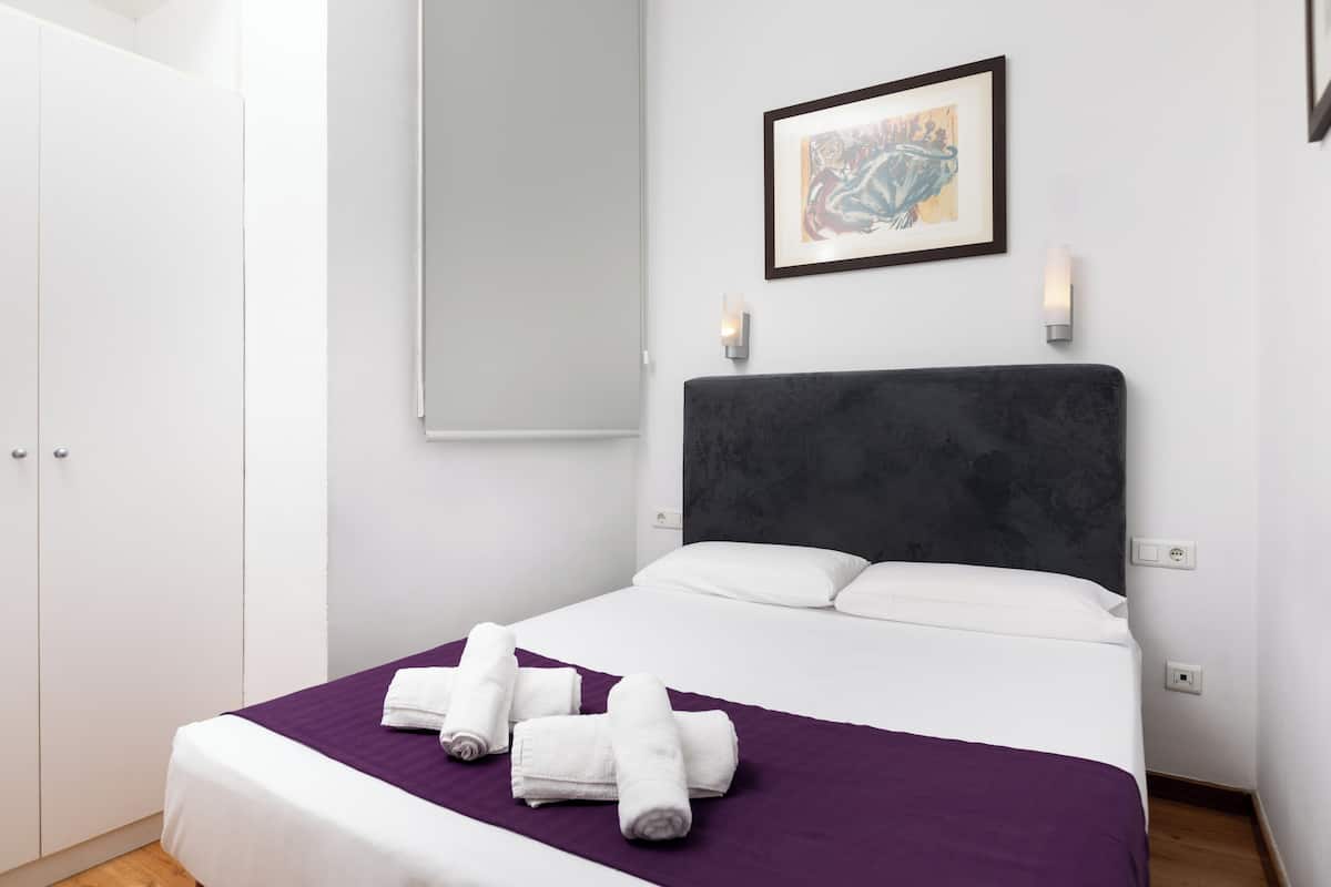 A neatly made bed with white linens and a purple blanket, a framed artwork on the wall, and a white wardrobe.