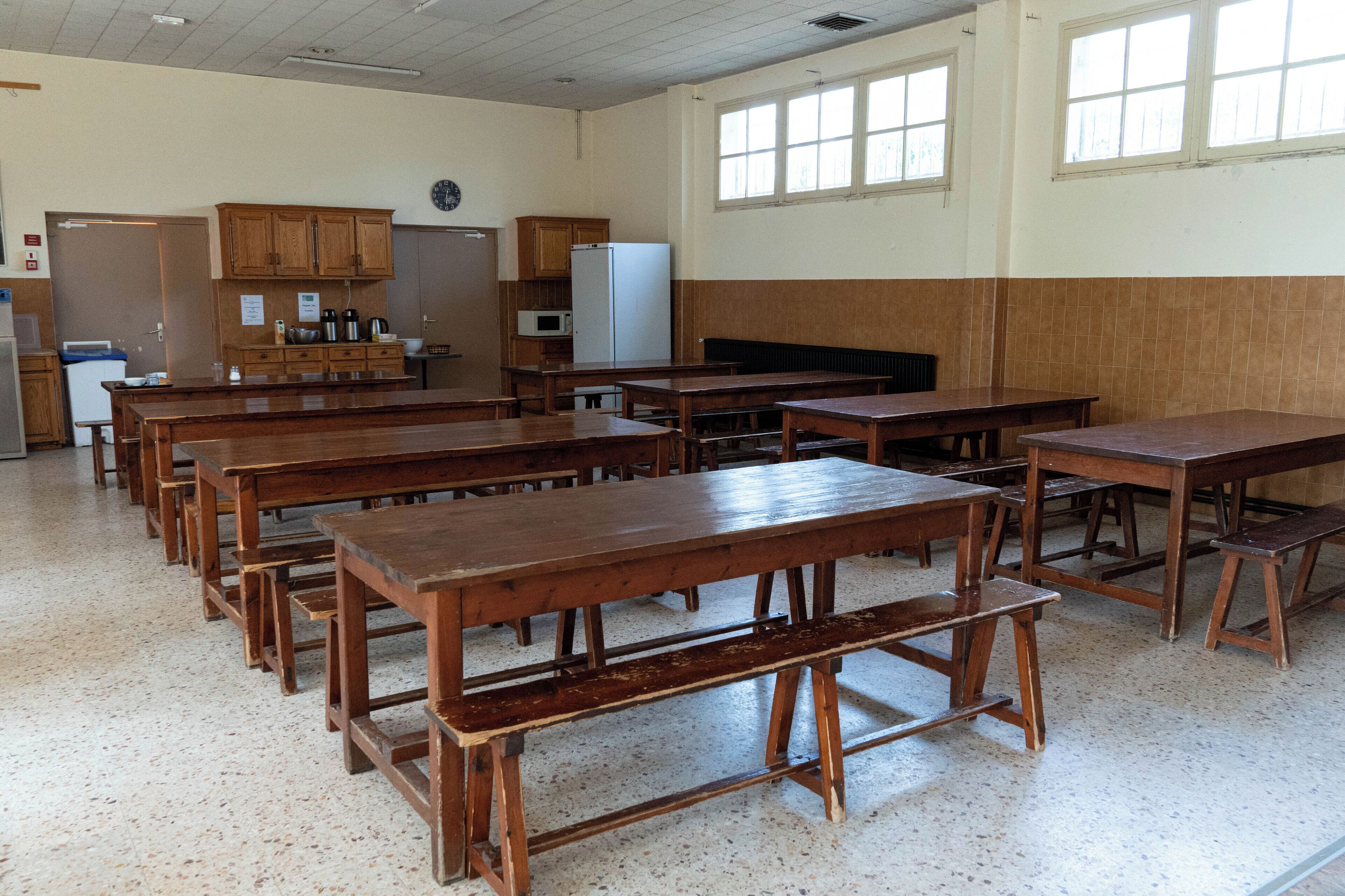 A classroom with wooden desks and benches, a kitchenette, and a clock on the wall.