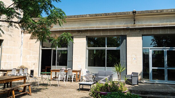 Un patio al aire libre con mesas y sillas, ventanas amplias y fachada de edificio de piedra.