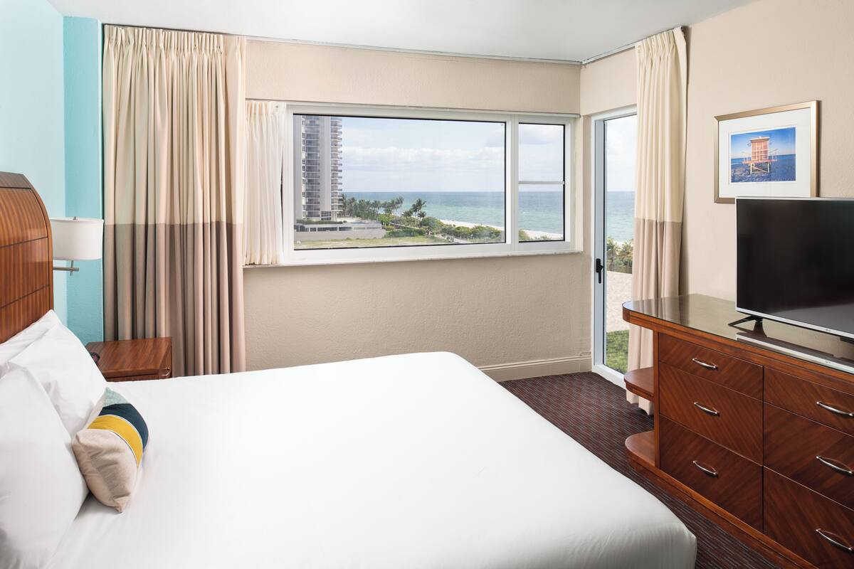 A hotel room with a large bed, a TV, a wooden dresser, and a window with a view of the ocean and a building.
