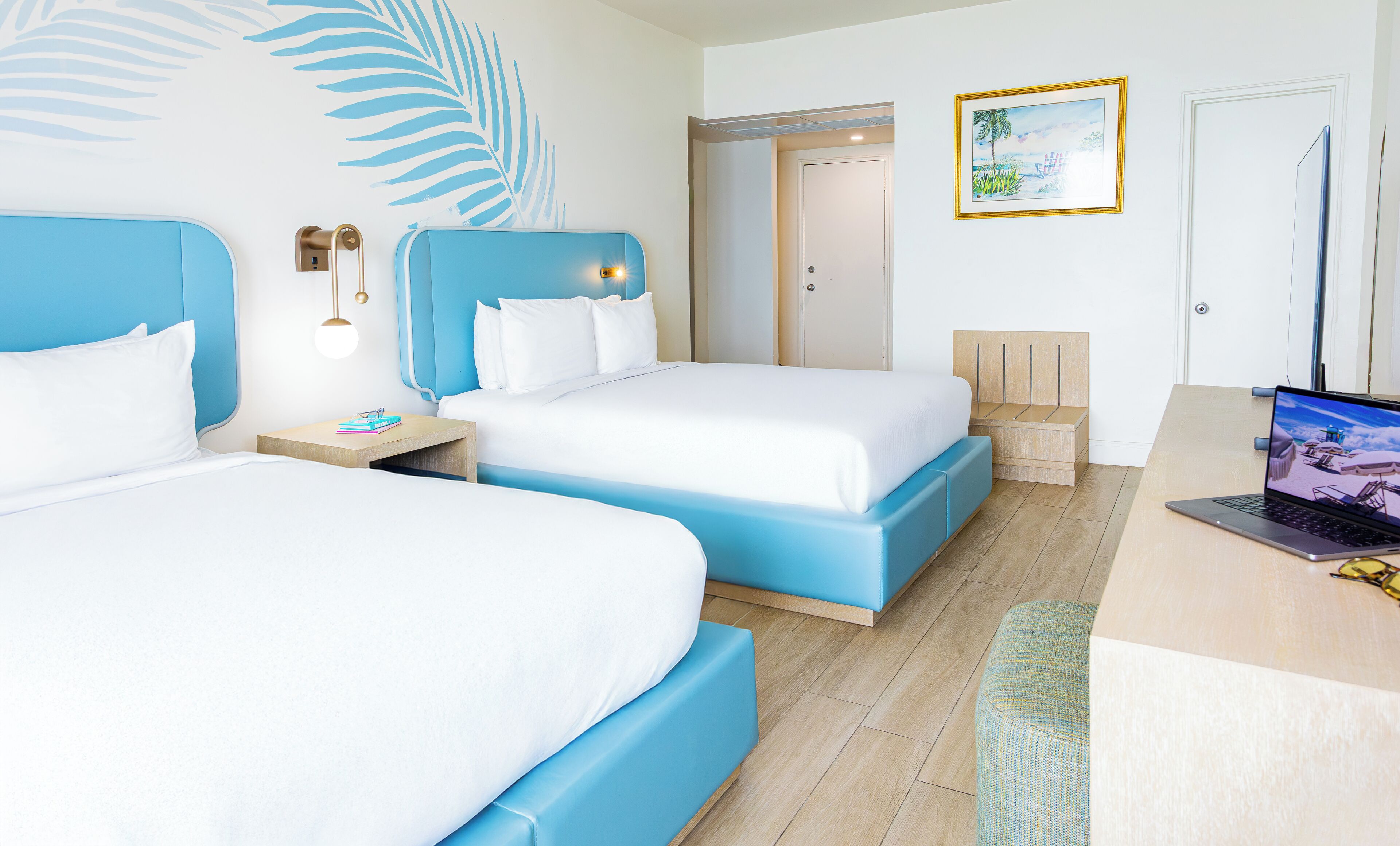 A hotel room with two beds, a desk with a laptop, a door, and a wall with a palm leaf design.