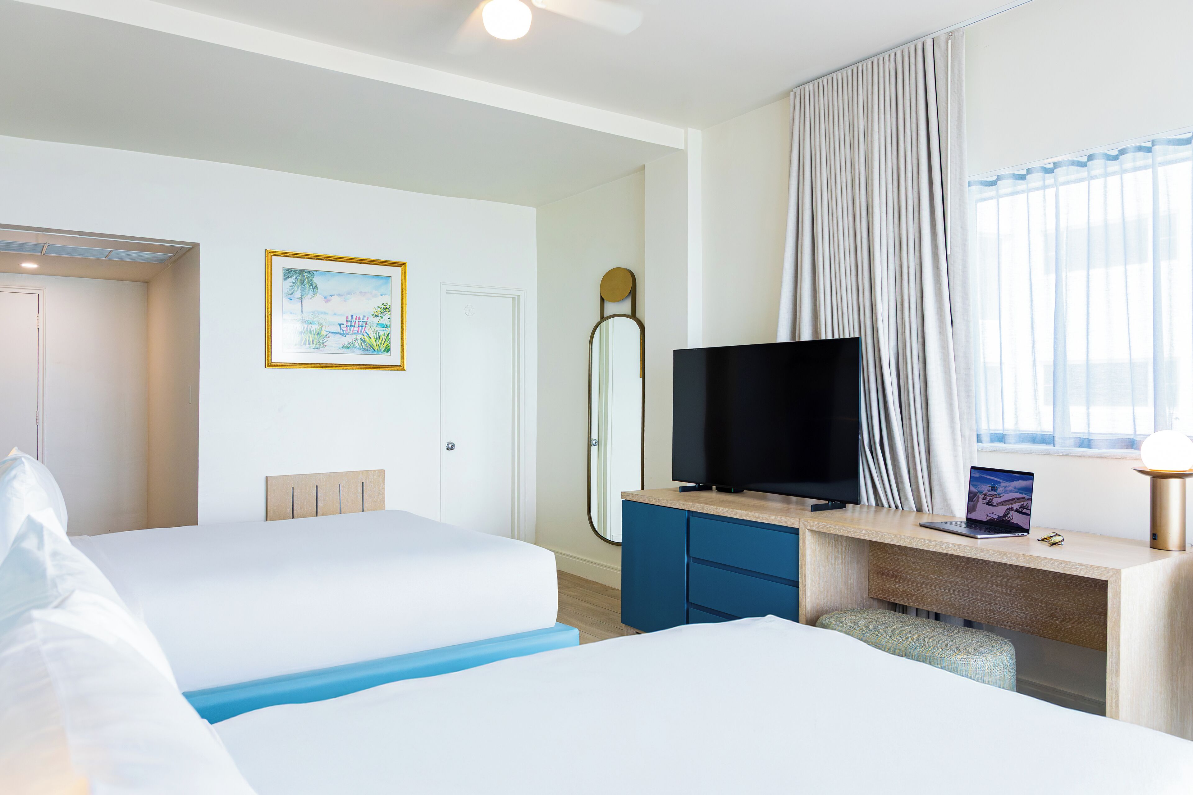 Een hotelkamer met twee bedden, een bureau met een laptop, een televisie en een groot raam met gordijnen.