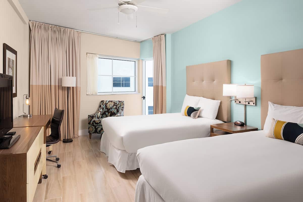 A hotel room with two beds, a desk, a chair, a lamp, and a window with curtains.