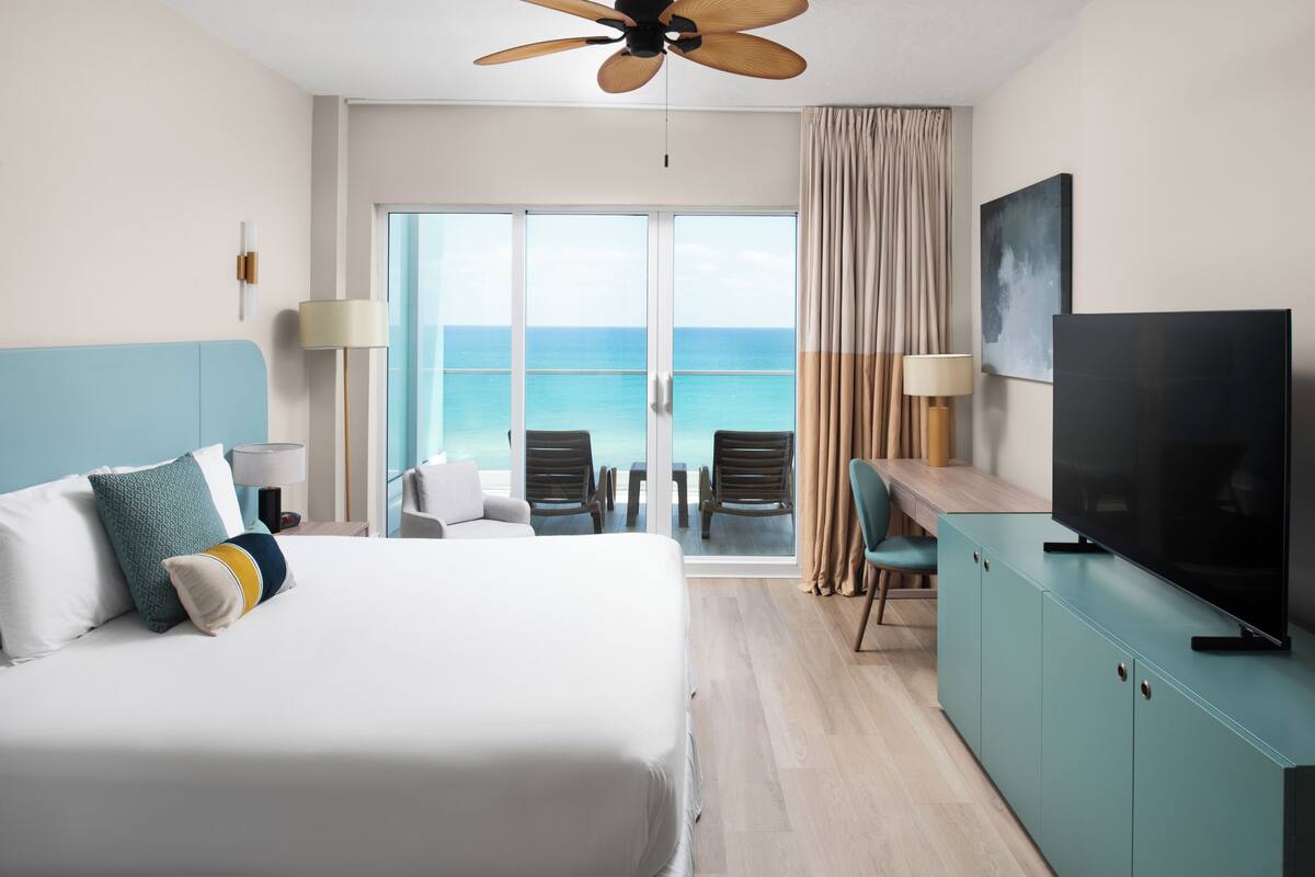 A hotel room with a large bed, a TV, a desk, and a view of the ocean.