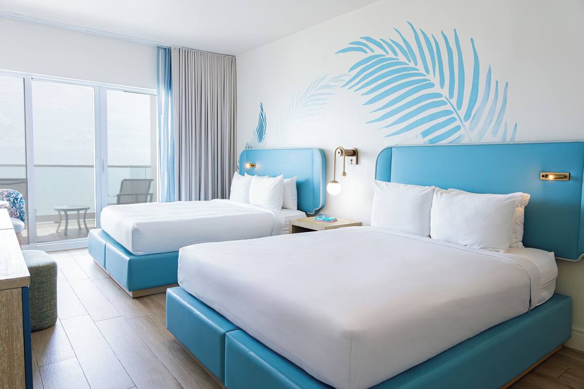 A hotel room with two beds, a large window with curtains, a small table, and a chair.