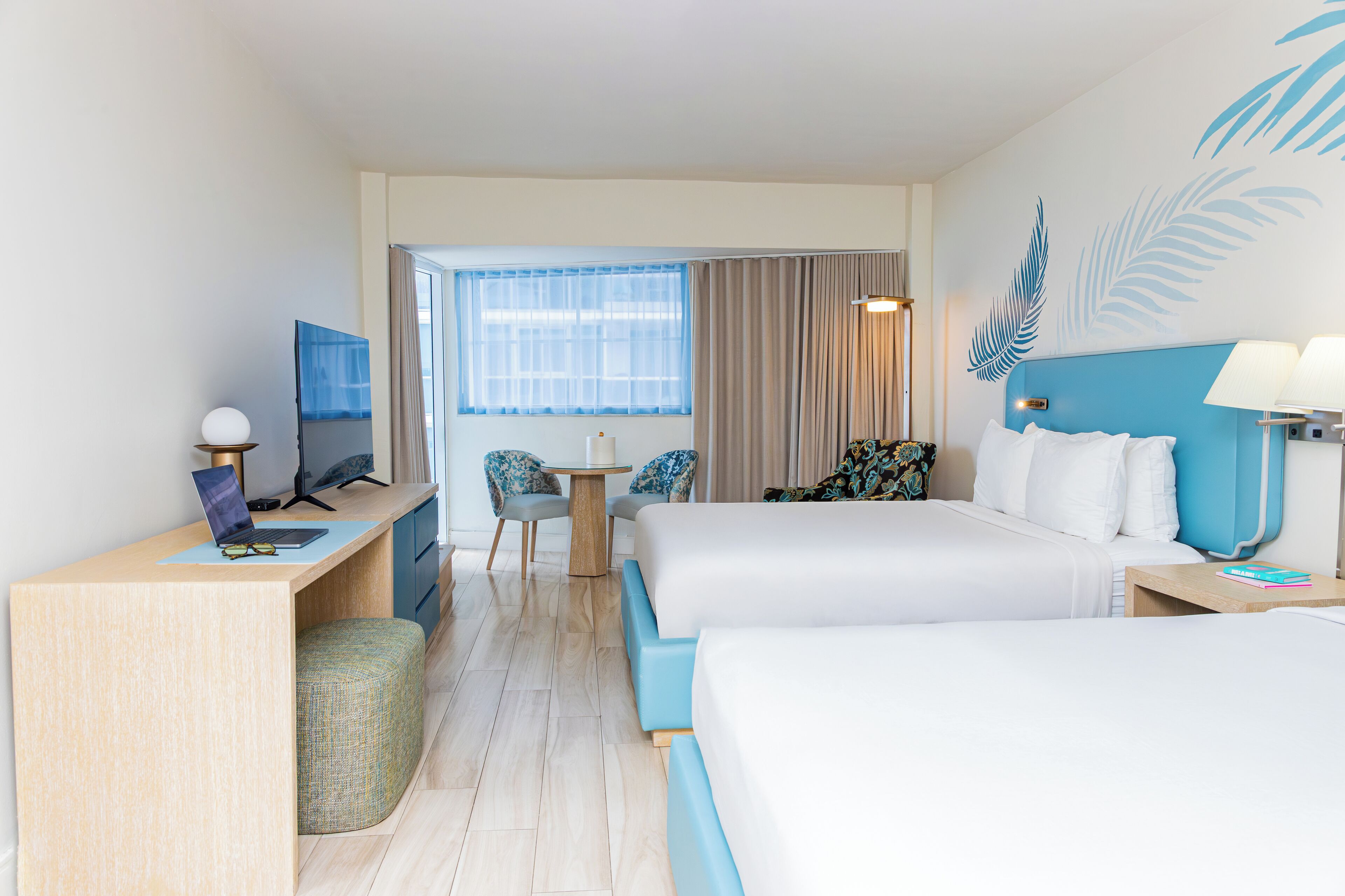 Habitación de hotel con escritorio, dos sillas, cama, mesita y ventana con cortinas.