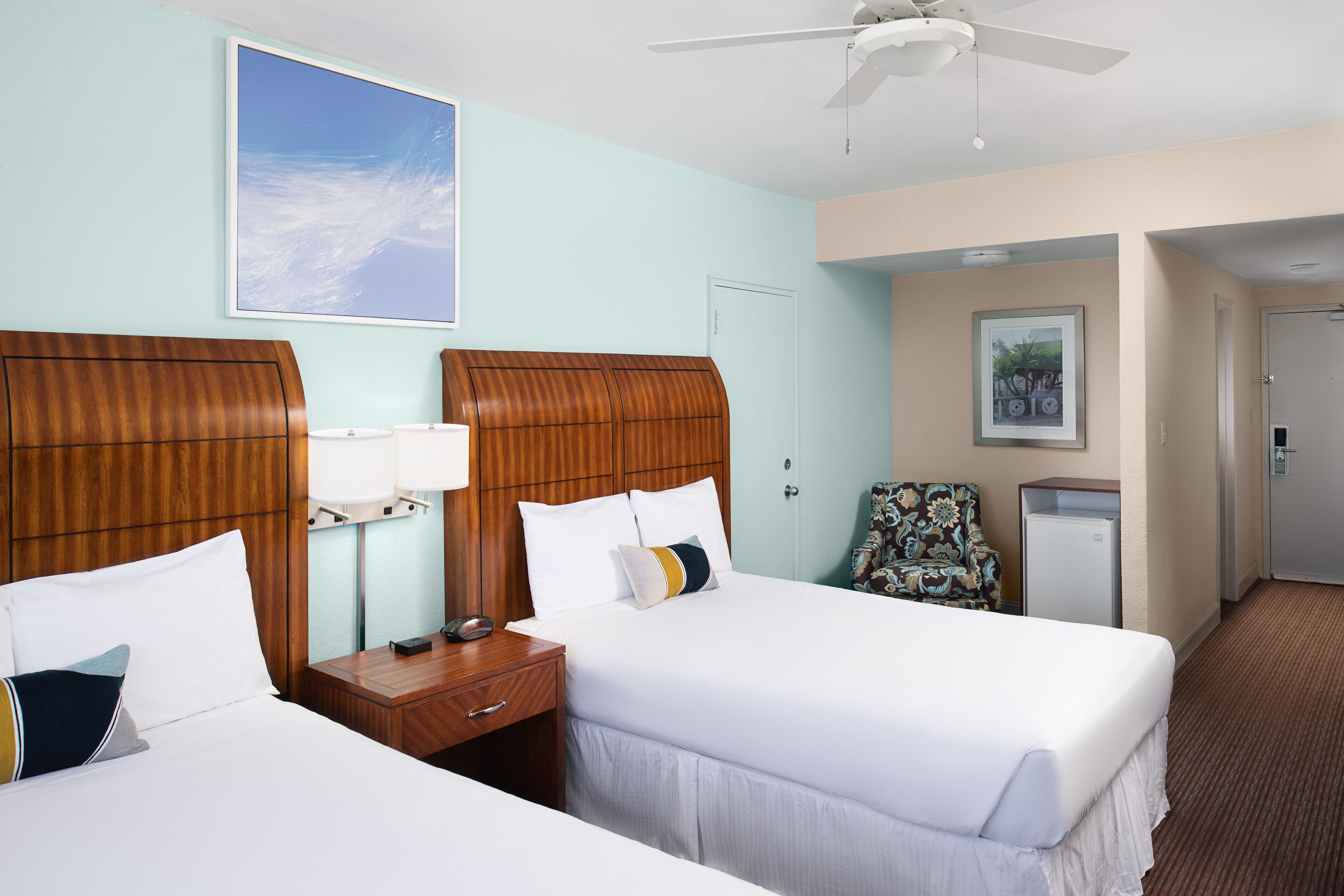 A hotel room with two beds, a ceiling fan, a desk with a lamp, a chair, a desk lamp, and a framed picture on the wall.