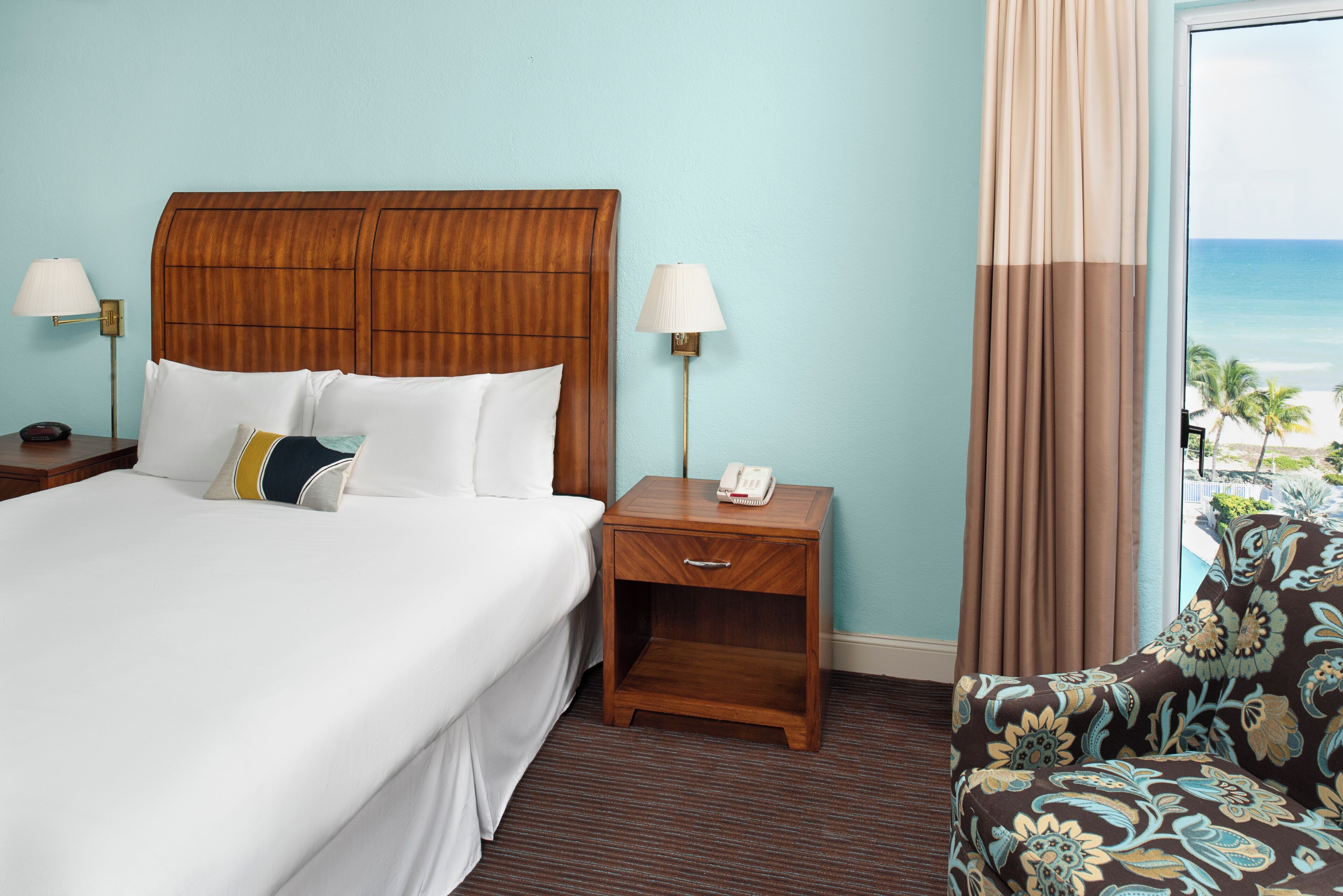A hotel room with a large bed, two bedside tables, a nightstand, a chair, and a view of the ocean.