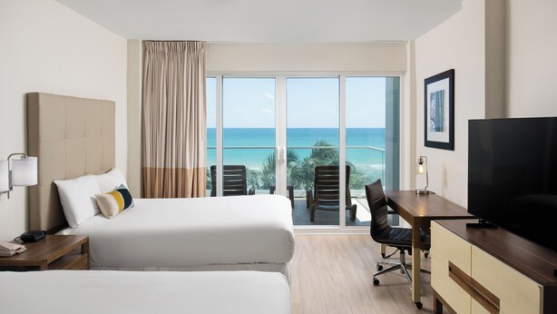 Habitación de hotel con dos camas, un escritorio, un televisor y un balcón con vista al océano.