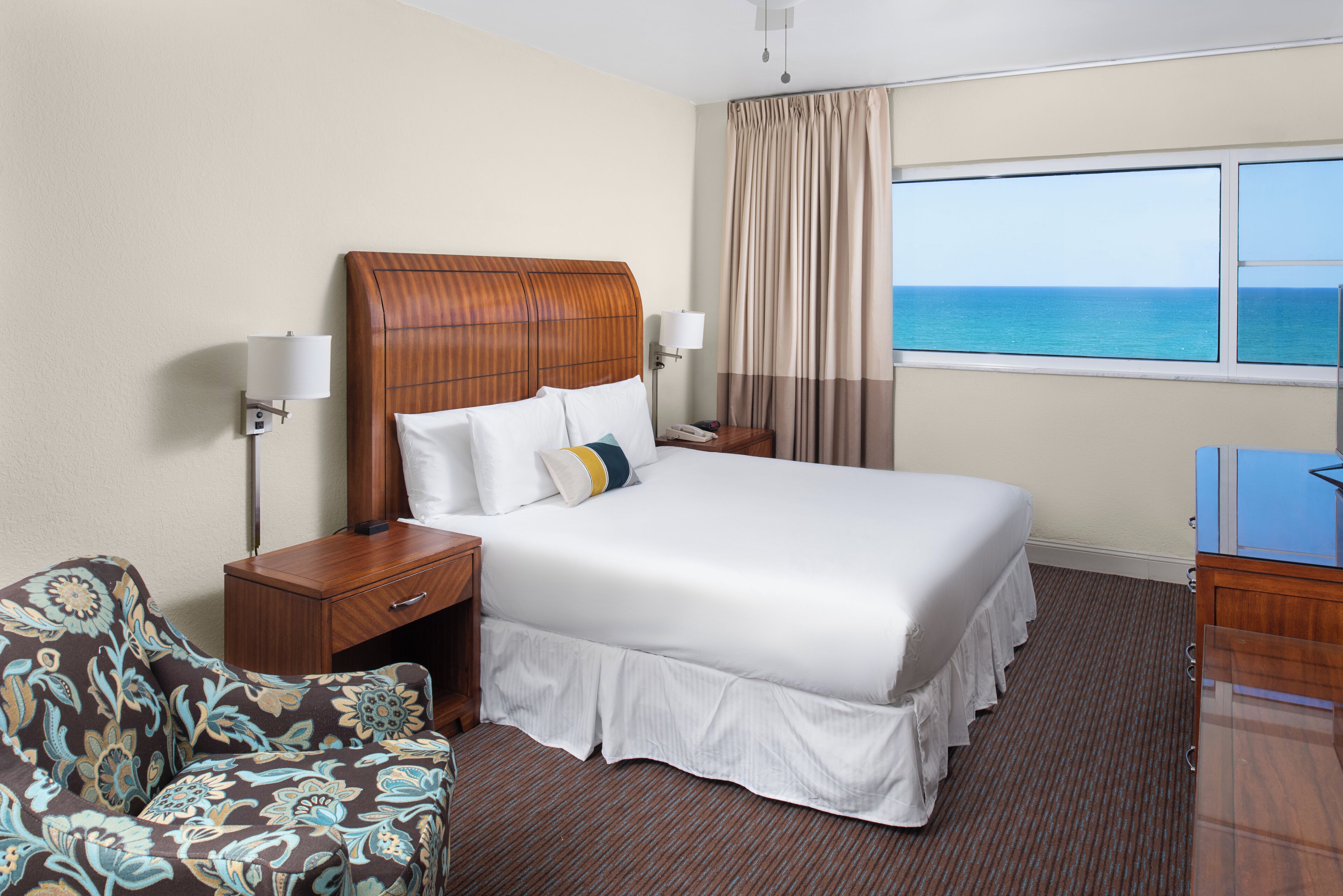 A hotel room with a large bed, a nightstand, a lamp, a chair, and a view of the ocean.
