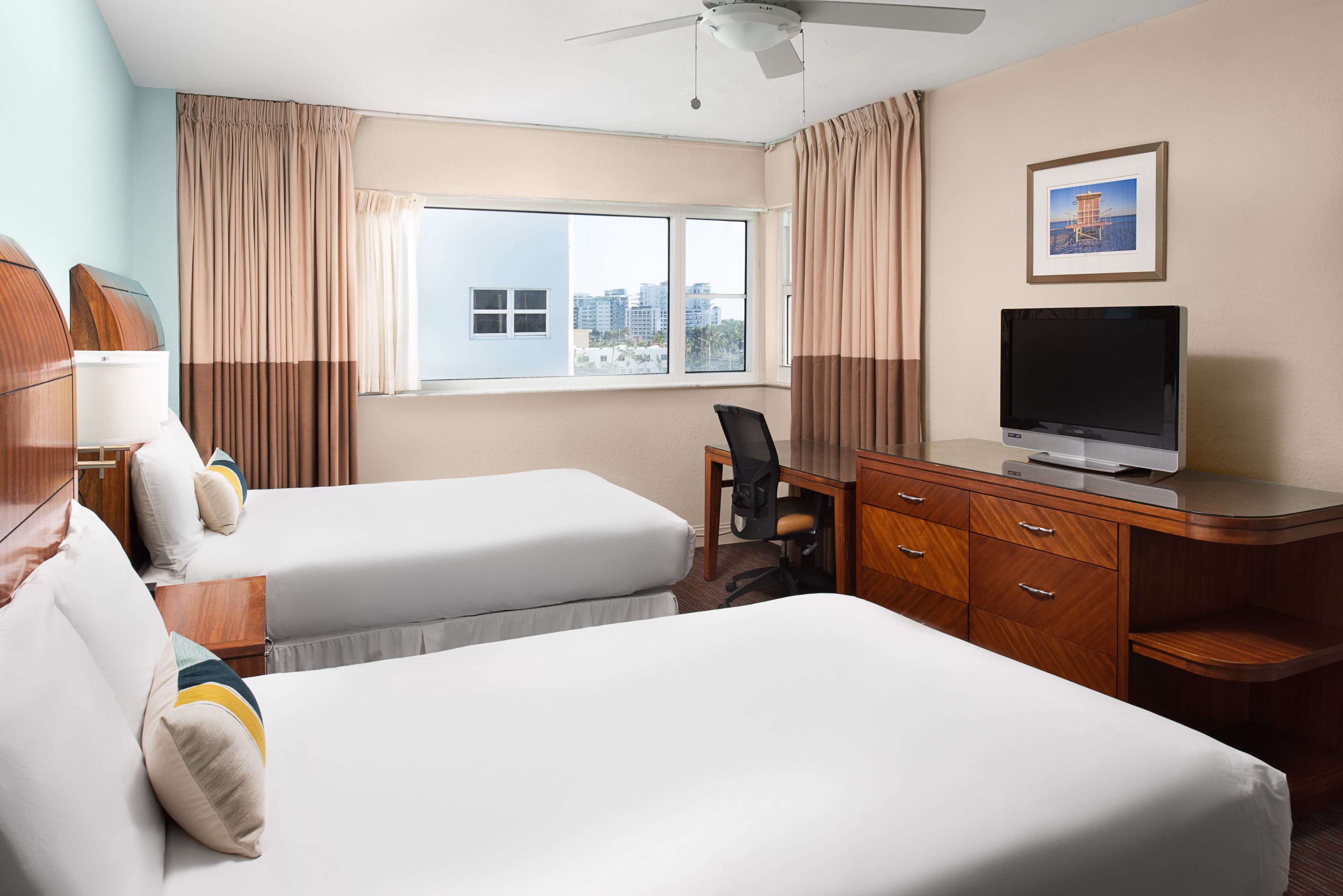 Quarto de hotel com duas camas, uma mesa de trabalho com uma televisão, uma cadeira e uma janela com cortinas.