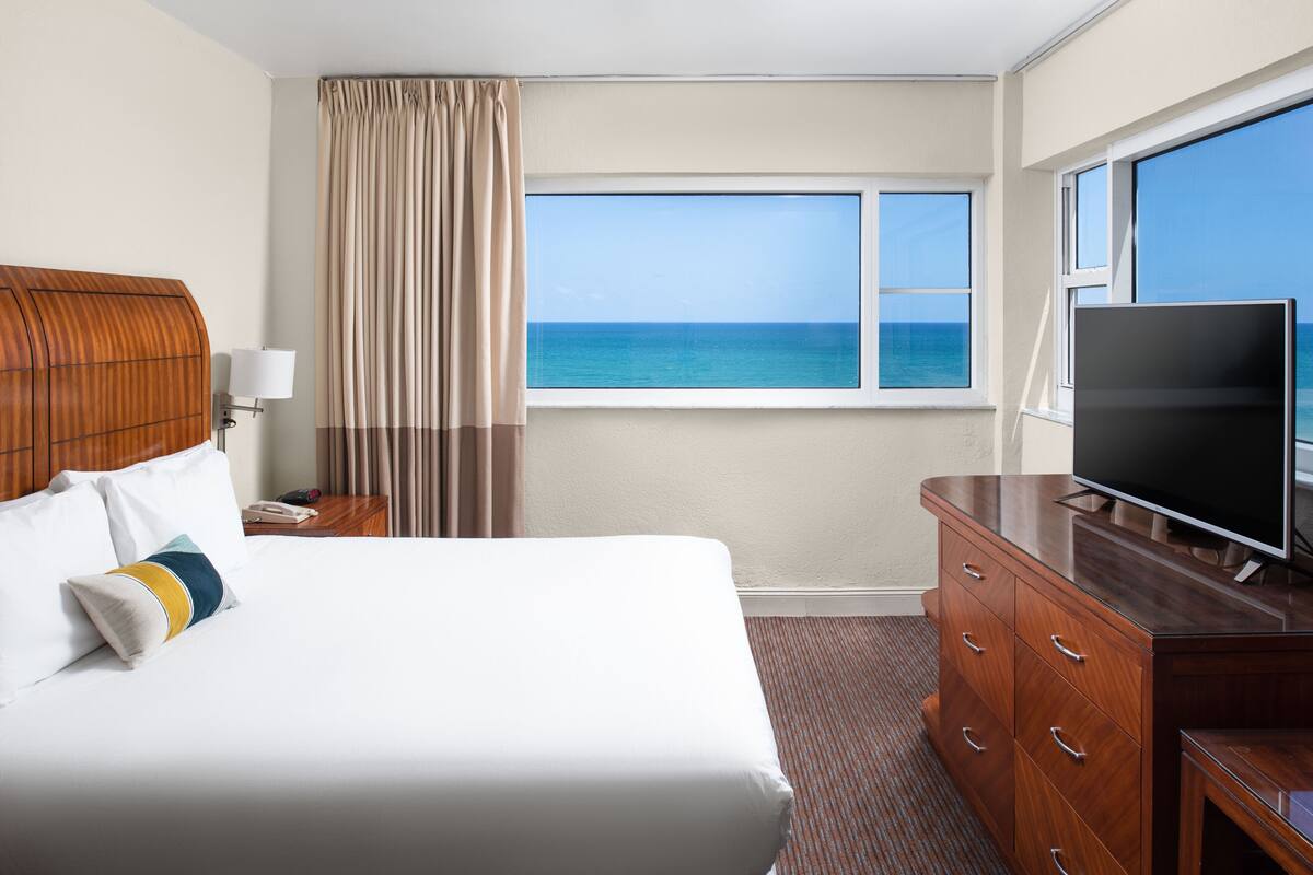 A hotel room with a large bed, a TV, a wooden dresser, and a window with a view of the ocean.