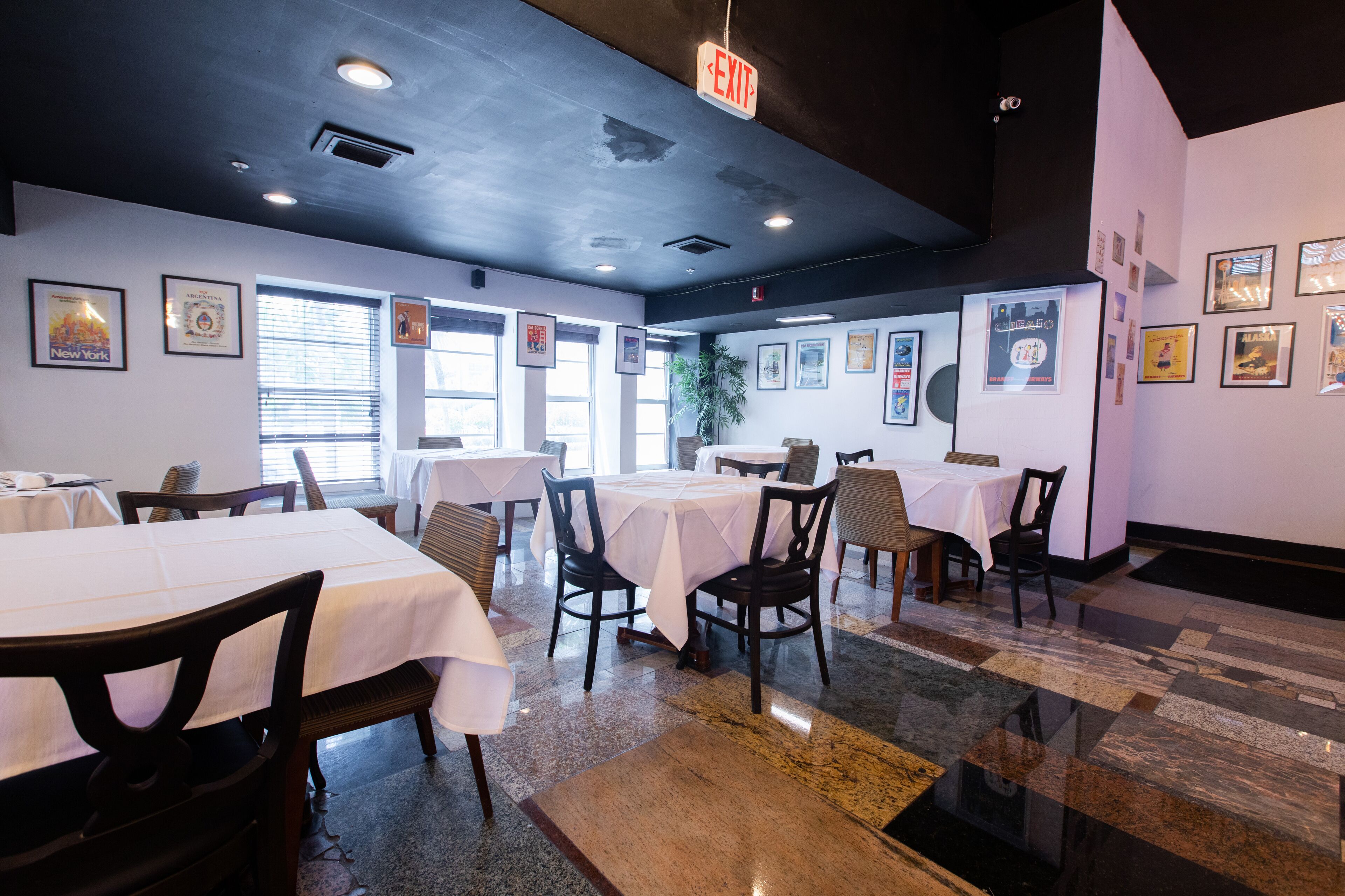 A restaurant with multiple tables and chairs, framed pictures on the walls, and an exit sign.