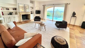 Un salón amplio con un sofá marrón, una mesa de centro y una televisión. Cuenta con una chimenea, una estantería y un gran ventanal con vistas al exterior.